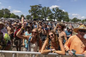 Large outdoor festival crowd behind a metal barrier on a sunny day, with a distant stage and trees.