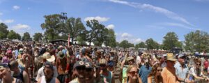Crowd at an outdoor music festival on a sunny day; people wear hats and sunglasses with tents in the background.