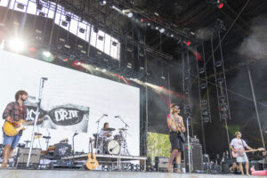 Band performing on an outdoor stage with guitars and drums and a large screen behind them.