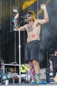Male punk singer shirtless on stage with microphone, beanie and sunglasses, arms painted in blue and red; text 'Cheap Folk Punk' on chest.