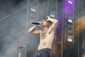 Male shirtless singer on stage, holding a microphone and singing, wearing sunglasses and jeans with stage fog and lighting in the background.