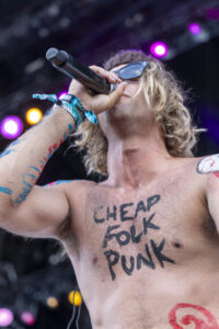 Male performer shirtless, painted arms, singing into a microphone on stage with colorful lights in the background.