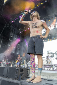 Shirtless rock singer on stage, body painted in bright colors, singing into a handheld microphone.