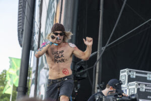 Shirtless punk singer on stage wearing a knit beanie and sunglasses, words 'CHEAP FOLK PUNK' on his chest while singing into a mic.