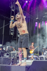 Shirtless male singer on stage, microphone in hand, arm raised amid purple stage lights and fog.