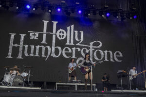 Band performs on a large stage with a black backdrop reading 'Humb...stone', lead singer with guitar center, blue stage lights above, drummer to left.
