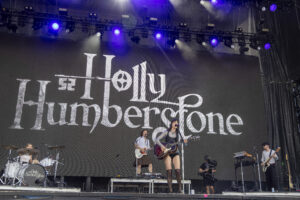 Live concert on a large stage; a giant backdrop reads 'Holly Humberstone' as the band performs.
