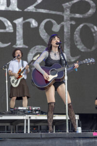 Female singer-songwriter in lace top sings into a microphone while playing a purple acoustic guitar on stage; a second guitarist is in the background.