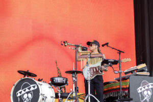 Musician playing trumpet at a stage with orange backdrop, wearing a cap and sunglasses, surrounded by drums and keyboards.