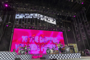 Live concert stage with a large pink video wall, musicians playing drums and guitar, and a black-and-white checkerboard stage front.