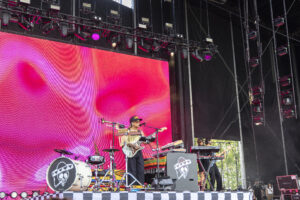 Musician wearing a hat and sunglasses performing on a stage with a bright pink LED backdrop and drum kit nearby.