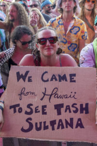 Smiling woman wearing sunglasses holds a large cardboard protest sign in a crowded outdoor event; sign reads 'WE CAME from Hawaii'.