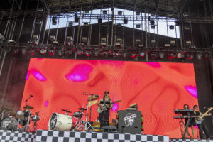 Live band performing on a large stage with a bright red LED screen backdrop, drums and guitars visible in the foreground.