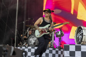 Female musician on stage playing a mint-green electric guitar, tattoos visible on arms, wearing a black tank top and cap with sunglasses.
