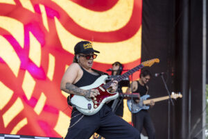 Female guitarist on stage playing a white electric guitar, wearing a black cap and sunglasses, tattooed arm, bright orange backdrop behind her.
