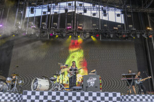 Live concert scene with a guitarist and band on a large stage, bright colorful lights, and a checkerboard front barrier while a drummer plays nearby.