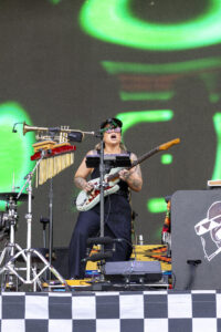 Female guitarist singing into a microphone on stage, wearing a cap and sunglasses, with a green abstract LED backdrop and assorted percussion/amp equipment nearby.