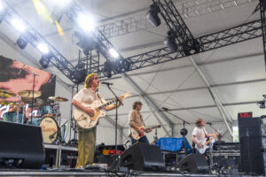 Band performs live on a stage with guitarists and a drummer under metal truss lighting rigs, mic stands nearby.