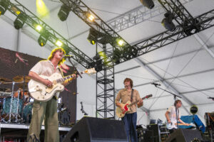 Live rock band performing on stage under metal truss with yellow lights; lead guitarist singing into a microphone.