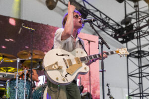 Male guitarist singing into microphone while playing a white hollow-body guitar on stage with drums in the background