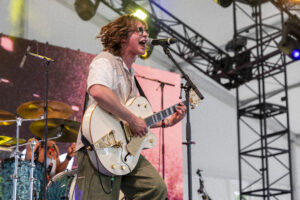 Musician with sunglasses singing into a microphone while playing a white electric guitar on stage, with a drummer in the background and stage rigging visible.