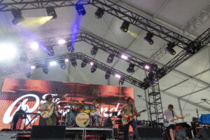 Band performs on a stage with multiple guitars, drums, and bright colored lights overhead on a large truss system.