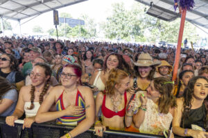 Crowd of festival-goers under a large canopy, many smiling, wearing colorful summer outfits in the front row near a barrier.