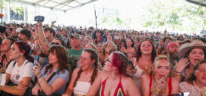 Crowd cheers at an outdoor concert under a large canopy, many people facing the stage and taking photos.
