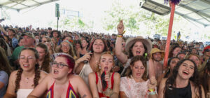 Large crowd at a summer festival under a canopy, many people cheering and dancing toward the stage.