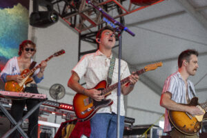 Male guitarist sings into mic while playing an orange-red electric guitar on a stage with other band members and equipment in the background, colorful lights overhead.