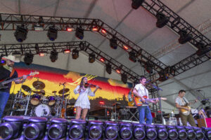Live concert on a large stage with musicians playing guitars and drums under a rig of metal trusses and red/blue lighting, foreground purple stage lights visible.