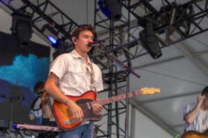 Male guitarist singing into a microphone on stage, playing a sunburst electric guitar with a strap, stage truss and blue lighting in the background