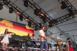 Band performs on a stage with multiple musicians under rigging and red stage lights, guitars and a keyboard visible in the foreground