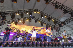 Band performs on a lit stage under metal truss rigging with guitars and drums visible