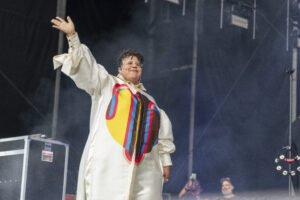 Performer on stage in a cream outfit with a bold colorful graphic, raising one arm to the crowd.