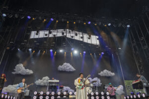 Live concert on a large outdoor stage with musicians playing guitars and keyboards beneath colorful spotlights and a cloud-themed backdrop.