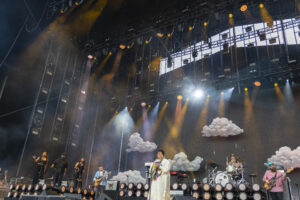 Stage view of a live band performing under bright spotlights, with fluffy cloud props on stage and a smoky atmosphere.