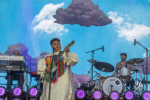 Guitarist in a colorful retro outfit plays a green electric guitar and sings into a mic on a bright stage with a blue sky backdrop and purple lights.