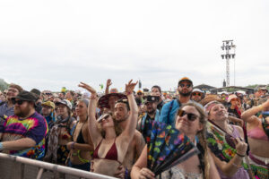 Crowd of festivalgoers wearing colorful outfits, raising hands at an outdoor concert barrier.