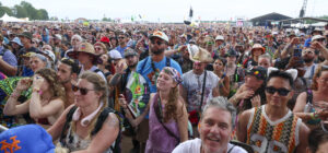 Large outdoor festival crowd with colorful outfits, hats, and sunglasses, facing a stage in the distance.