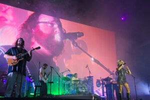 Guitarist with beard plays an electric guitar on a smoky, colorful concert stage with a singer at right and a large screen backdrop behind them.