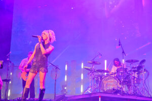 Female singer in a sparkly dress singing into a microphone on a purple-lit stage with band in background.