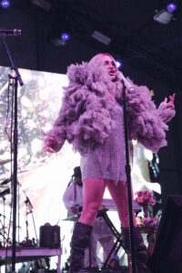 Female singer on stage wearing a large lavender feather coat and a glittery dress, singing into a microphone under purple stage lights.