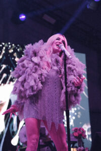 Female singer on stage wearing a pink feather boa, singing into a microphone under purple lighting.