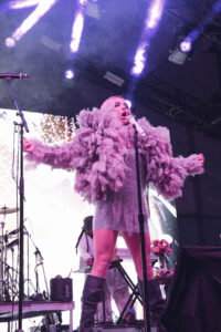 Singer on stage wearing a pink feathered costume, singing into a microphone amid purple stage lights.