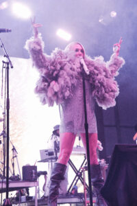 Singer on stage wearing a fluffy feathered jacket, holding a microphone, raising both hands in a performance moment.