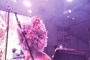 Female singer on stage wearing a pink feathered outfit, holding a microphone, performing under purple stage lighting.