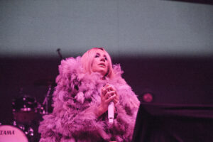 Singer on stage bathed in pink light, wearing a pink feathered cape and holding a microphone.