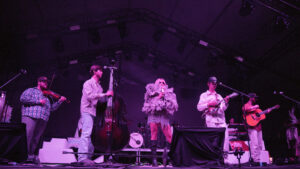 Band performs on a purple-lit stage: center singer in a fluffy feathered outfit with a microphone, surrounded by violinist, upright bassist, guitarist, and other musicians.