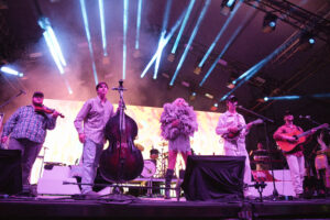 Live concert on a large stage with blue spotlights; center-front singer in a fluffy feathered outfit, surrounded by musicians playing violin, upright bass, and guitars while drums rise in the back.